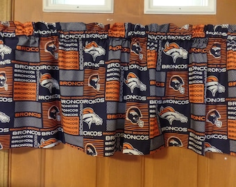 May include: A fabric window valance featuring a repeating pattern of the Denver Broncos logo and the word Broncos in orange and white on a dark blue background. The valance has a gathered rod pocket top for hanging and a pleated, ruffled design.
