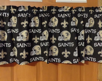 May include: Black and gold fabric valance with a repeating pattern of New Orleans Saints football helmets and fleur-de-lis. The valance has a ruffled edge.