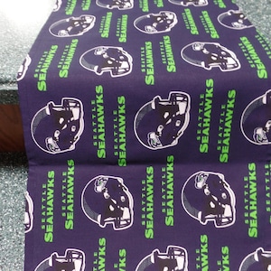 May include: A navy blue table runner featuring a repeating pattern of Seattle Seahawks football helmets and team name in white and green. The runner is made of fabric.
