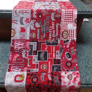 May include: A rectangular fabric table runner featuring a patchwork design with various Ohio State University themed prints in red, white, gray, and black. The pattern includes the Brutus Buckeye mascot, footballs, helmets, and text reading Ohio State.