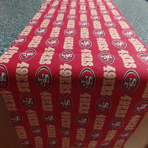 May include: Red table runner with a repeating pattern of the San Francisco 49ers logo in gold. The logo includes the team's name and the number 49.