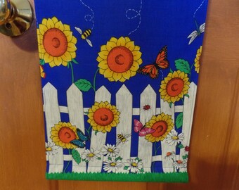 May include: A blue fabric kitchen towel with a white picket fence design. The fence is decorated with sunflowers, butterflies, bees, and daisies.