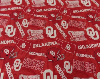 May include: Red fabric with a white repeated pattern of the Oklahoma Sooners logo, including the words "Oklahoma", "Sooners", and "Boomer Sooner".