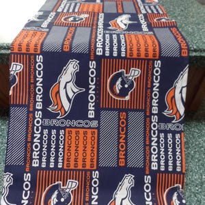 May include: A navy blue table runner featuring a repeating pattern of the Denver Broncos logo, helmet, and the word "BRONCOS" in orange and white. The runner is made of fabric and is suitable for a dining table or other surfaces.