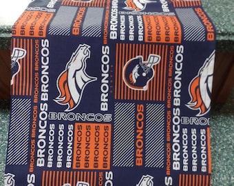 May include: A navy blue table runner featuring a repeating pattern of the Denver Broncos logo, helmet, and the word "BRONCOS" in orange and white. The runner is made of fabric and is suitable for a dining table or other surfaces.