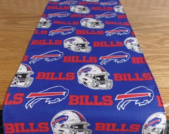 May include: A rectangular table runner in royal blue featuring a repeating pattern of white football helmets with red and blue details, the red text "BILLS", and the red and blue Buffalo Bills team logo printed across the fabric surface.