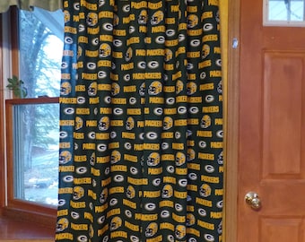 May include: Green and yellow Packers-themed curtain panel. The fabric features a repeating pattern of the team's logo, helmets, and the word "Packers" in a bold font. The curtain is hanging in front of a window and a wooden door.