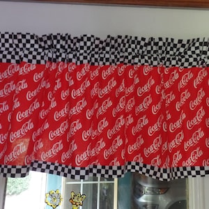 May include: A red window valance featuring the Coca-Cola logo in white script. The valance has a black and white checkered pattern at the top and bottom edges. The valance is hanging on a white rod.
