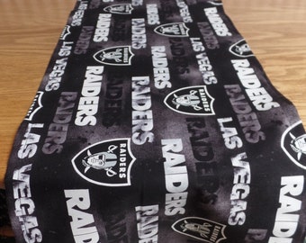 May include: Black and white table runner with a repeated pattern of the Las Vegas Raiders logo and the words "Las Vegas Raiders".