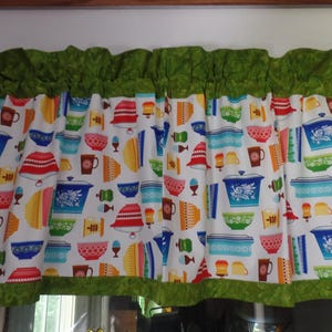 May include: A kitchen valance with a white background, featuring a colorful pattern of kitchenware, including bowls, pots, and mugs. The valance has a green ruffled top and bottom trim, adding a decorative touch to the window treatment.