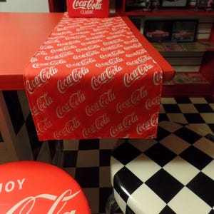 May include: A red table runner featuring a repeating white Coca-Cola logo pattern, positioned on a red surface with a six-pack of glass Coca-Cola Classic bottles in a red cardboard carrier placed at the end of the runner.