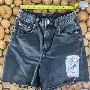 May include: Black denim shorts with a raw hem and a decorative patch. The shorts measure approximately 10 inches across the waist. The patch is white with a red and black design.
