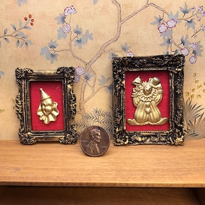 May include: Two gold framed pictures with red velvet mats. The picture on the left features a gold clown head. The picture on the right features a gold clown figure.