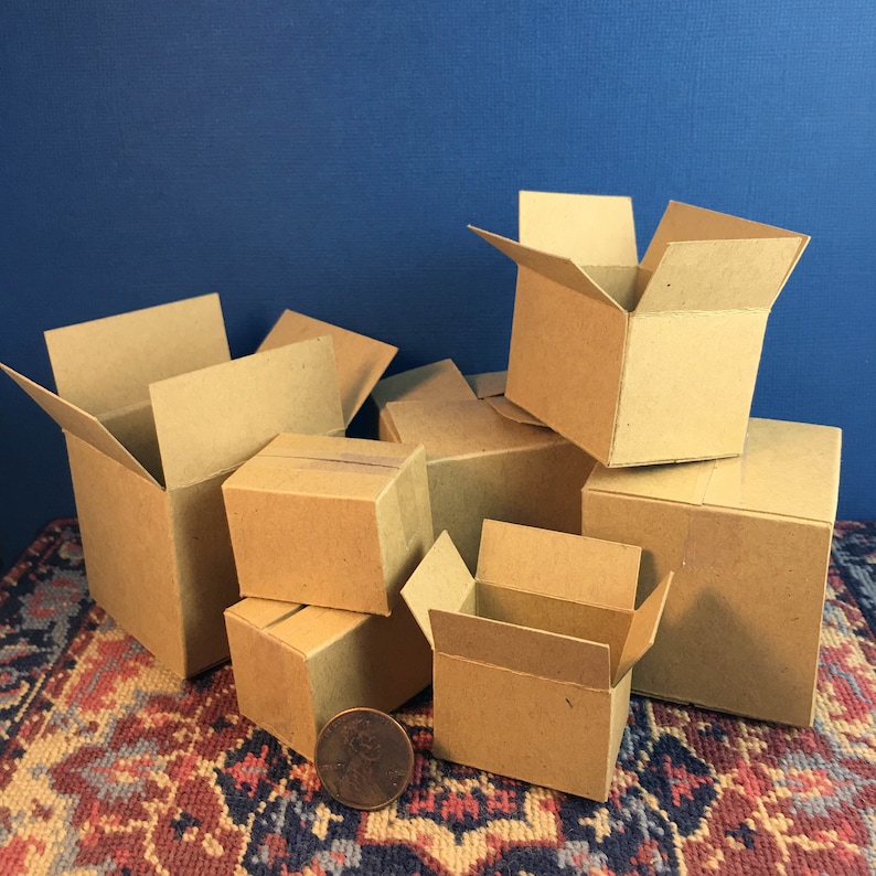 May include: A collection of small, open cardboard boxes in various sizes, some stacked and others with flaps open. A penny is placed near the boxes for scale. The boxes are on a patterned rug with a blue background.