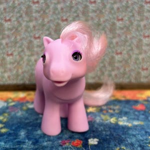 Puede incluir: Un poni de juguete rosa claro con melena y cola rosas. El poni tiene ojos morados y está sobre una superficie azul con estampado floral. El fondo es un papel pintado con dibujos.