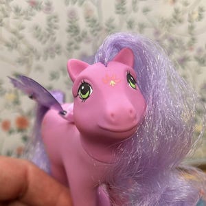 Peut inclure: Une figurine de poney rose avec de longs cheveux lavande fluides et de grands yeux verts. Le poney présente un petit symbole de fleur rose et jaune stylisé sur le front et des ailes violettes translucides attachées au dos, en plastique.