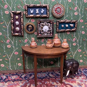 Puede incluir: Una escena de habitación en miniatura con arte de conchas marinas enmarcadas, cerámica y una mesa de madera. La pared está decorada con obras de arte enmarcadas y papel pintado floral. Una pequeña figura de elefante de madera está en el suelo. La mesa contiene varias piezas de cerámica marrón y naranja.