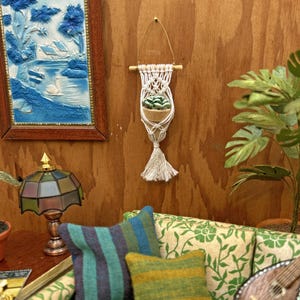 May include: A miniature white macrame plant hanger suspended from a thin gold wire and a wooden dowel. The hanger holds a small tan pot containing a green succulent. The piece features intricate knotted cord patterns and a long fringe at the base.