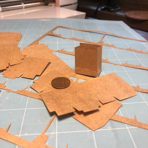 May include: A miniature brown paper bag stands upright amidst scattered cut-out cardboard pieces and a penny on a blue cutting mat. The bag is rectangular with a folded top, suggesting a craft project or packaging design.