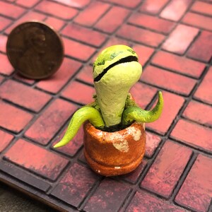 May include: A small green and brown ceramic plant sculpture in a terracotta pot. The plant has a long, thin stem with a rounded top and a few small leaves.