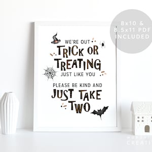 May include: A framed Halloween-themed print with the text "We're out Trick or Treating just like you, please be kind and just take two." The print includes a witch's hat, spider, web, and bat illustrations. A white ceramic house and vases are in the foreground.