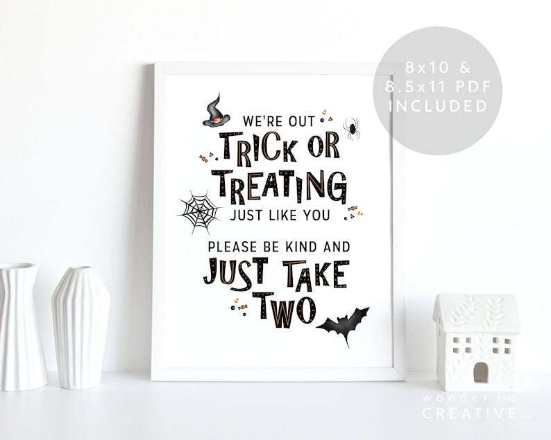 May include: A framed Halloween-themed print with the text "We're out Trick or Treating just like you, please be kind and just take two." The print includes a witch's hat, spider, web, and bat illustrations. A white ceramic house and vases are in the foreground.