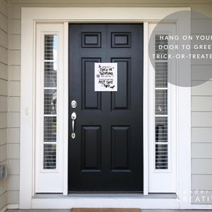 May include: A black front door with a white framed window on each side. A white sign with black text that reads "Trick or Treating, Just Take Two" is hanging on the door. The text "Hang on your door to greet trick-or-treaters" is also visible.
