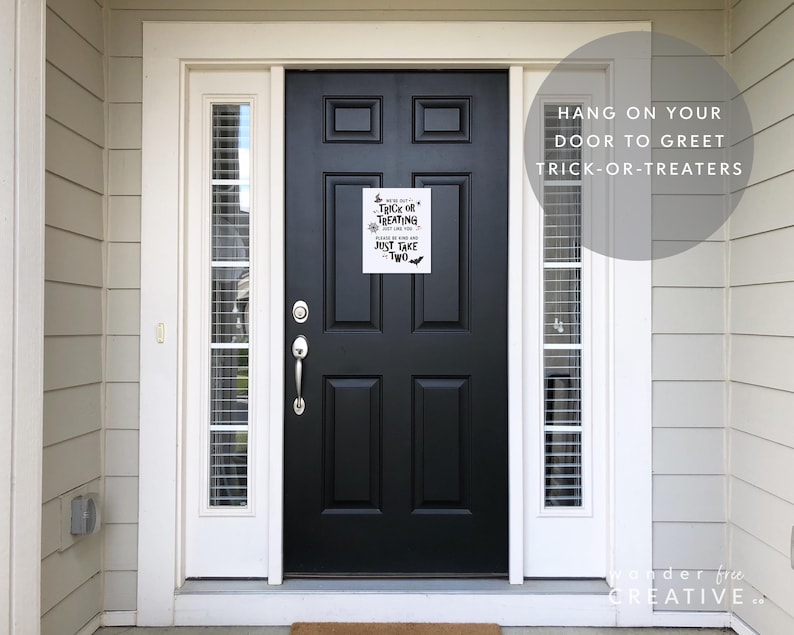 May include: A black front door with a white framed window on each side. A white sign with black text that reads "Trick or Treating, Just Take Two" is hanging on the door. The text "Hang on your door to greet trick-or-treaters" is also visible.
