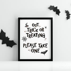 May include: A framed Halloween sign with the words "Out Trick or Treating Please Take One" in black lettering. The sign is decorated with a witch's hat, spider, web, and bat illustrations. Black bat cutouts are on the wall.