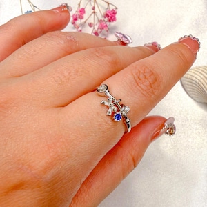 May include: A silver ring featuring a bow, a heart, and a blue gemstone. The ring has a delicate design with small white gemstones. The ring is on a finger with manicured nails.