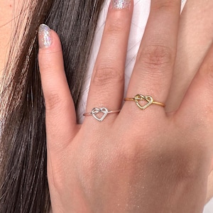May include: Two heart-shaped rings, one silver and one gold, are displayed on a hand. The rings have a delicate, intertwined design forming a heart shape. The gold ring has a shiny finish, while the silver ring has a matte appearance.