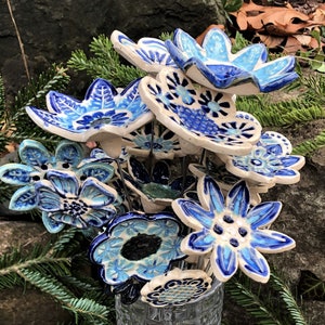 May include: A display of ceramic flowers in a clear glass vase. The flowers are in shades of blue and white, with detailed patterns. The vase is on a stone surface, surrounded by evergreen branches.