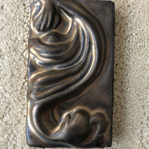 May include: A ceramic tile with a raised design of a swirling, abstract shape. The tile is a dark brown color with a metallic gold sheen.