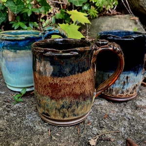 May include: Three ceramic mugs with textured handles and glazed finishes. One mug features a blue and teal gradient, another has a brown and tan drip pattern, and the third displays a deep black and blue ombre effect with a glossy surface.