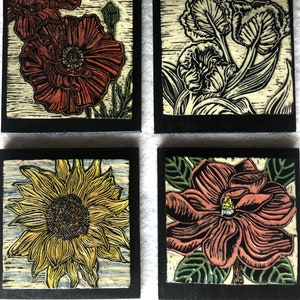 May include: Four square ceramic tiles with black frames, each featuring a different flower design. The top left tile shows a red poppy, the top right tile shows a white tulip, the bottom left tile shows a yellow sunflower, and the bottom right tile shows a pink magnolia.