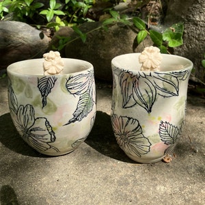 May include: Two white ceramic mugs with a floral design. The mugs have a green and pink floral pattern with black outlines. Each mug has a small white ceramic flower on the rim.