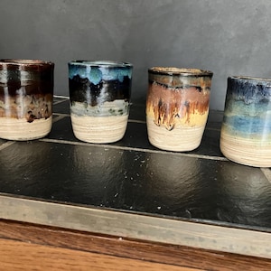 May include: Four handmade ceramic tumblers with a rustic, glazed finish. Each tumbler features unique color gradients, including shades of brown, blue, and yellow, with a natural, unglazed base. The tumblers are arranged on a dark, tiled surface.