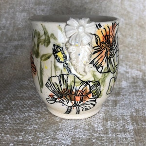 May include: A ceramic mug with a cream-colored base, adorned with hand-painted orange and black poppy flowers and green foliage. A white, three-dimensional flower is attached to the handle. The mug is approximately 4 inches tall.