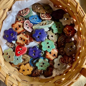 May include: A collection of colorful ceramic buttons in various shapes and sizes, including flowers, hearts, and leaves. The buttons are arranged in a wicker basket.