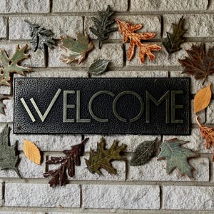 May include: A black metal welcome sign with the word "WELCOME" in silver lettering, surrounded by decorative ceramic leaves in shades of green, brown, and orange. The sign is mounted on a light gray brick wall.