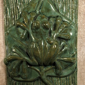 Könnte beinhalten: Eine grüne Keramikfliese mit einer in Relief geformten Frosch. Der Frosch sitzt auf einem Blatt mit ausgestreckten Beinen. Die Fliese hat eine strukturierte Oberfläche.