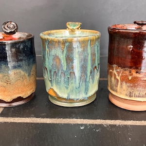 Pode incluir: Três potes de cerâmica feitos à mão com tampas. Os potes têm uma aparência rústica com uma variedade de cores, incluindo azul, marrom e verde. Cada pote tem um design de tampa único, adicionando ao seu apelo artístico. Estes potes são ideais para armazenamento na cozinha.