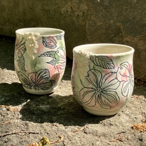 May include: Two white ceramic mugs with a floral design. The mugs have a pink and green watercolour background with black floral outlines. The mugs have a 3D floral design on the side.
