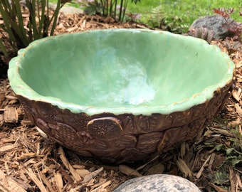May include: A green ceramic bird bath with a textured brown exterior. The bird bath is sitting on a bed of wood chips.