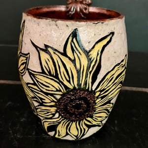 May include: A ceramic mug with a speckled off-white exterior featuring a carved and painted yellow sunflower with a dark brown center. The interior of the mug is a deep reddish-brown, and the handle is shaped like a textured brown leaf.