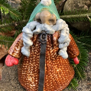 May include: A ceramic gnome figurine with a brown coat, red mittens, and a green hat. The gnome has a white beard and a black zipper down the front of the coat. The figurine is set against a backdrop of green pine needles and a stone.