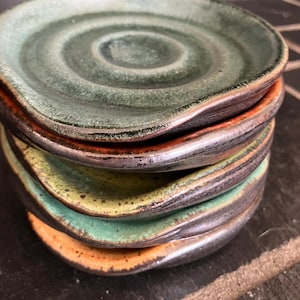 Könnte beinhalten: Ein Stapel von vier handgefertigten Keramik-Löffelablagen. Jede Ablage hat eine andere Farbe: Grün, Gelbgrün, Türkis und Orange. Die Ränder sind dunkelbraun. Die Löffelablagen haben eine kreisförmige Vertiefung in der Mitte.