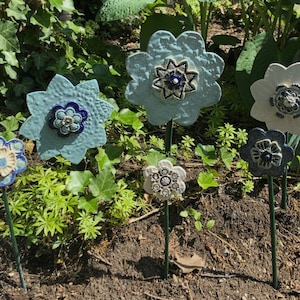 Pode incluir: Cinco esculturas de flores de cerâmica em cores azul, branco e preto com desenhos intrincados. As flores estão em estacas de metal e são colocadas num jardim.