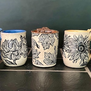 May include: Five ceramic mugs arranged in a row, each featuring a cream-colored body with black botanical illustrations. The mugs have distinct colored handles and interior glazes in red, blue, brown, yellow, and pink, with sculpted floral details.