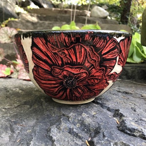 May include: A white ceramic bowl with a black rim and red poppy flower design. The bowl is sitting on a gray rock surface.